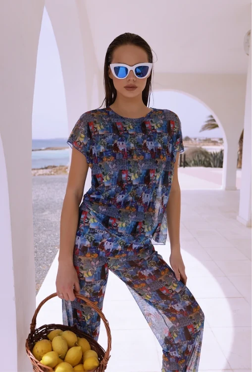 A model in white sunglasses poses outdoors under arches wearing the Mediterranean collage sheer blouse and matching pants. A basket of lemons is in her hand, and the sea is visible in the background.
